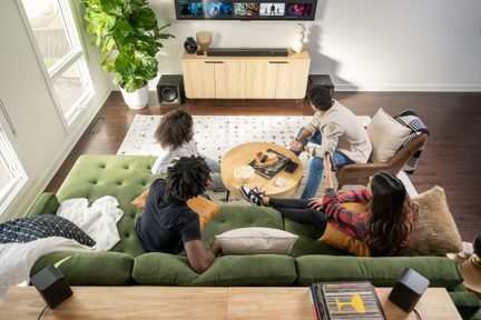 Quatre personnes assises sur un canapé vert regardant un écran de télévision, dans un salon avec un tapis et des plantes