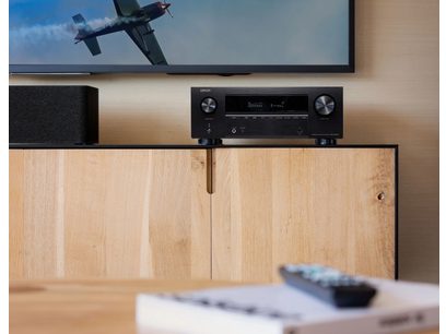 Ampli home-cinéma Denon AVR-X2800H DAB Noir, vue de face, télécommande sur meuble bois clair, avion militaire sur écran TV