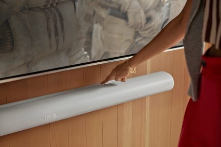 Barre de son blanche sur mur en bois, écran au-dessus, main droite pointant vers le haut de la barre