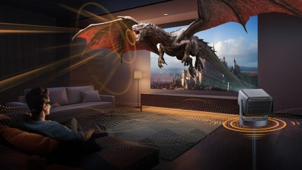 Salon sombre, canapé gris, lampe sur pied, projecteur, écran avec dragon volant, personne assise avec lunettes 3D