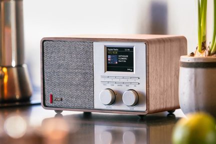 Radio numérique compacte en bois clair, façade aluminium brossé avec grille de haut-parleur en tissu gris sur la gauche, écran couleur, cinq petits boutons et deux gros boutons ronds, vue de trois-quarts avant