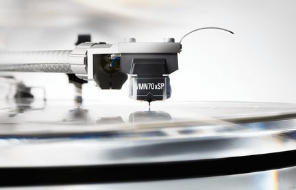 Zbliżenie z boku na czarno-szarą wkładkę gramofonową z plastiku i metalu zamocowaną na srebrnym ramieniu, z igłą dotykającą błyszczącej powierzchni płyty winylowej