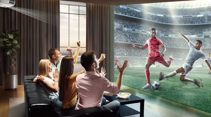 Vy bakifrån i trekvartsvinkel av ett modernt vardagsrum, vit projektor i taket som projicerar en fotbollsmatch på en stor skärm framför en svart hörnsoffa i läder där flera entusiastiska åskådare sitter