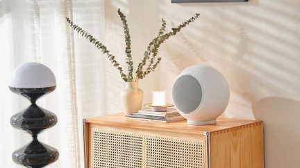 Ensemble décoratif intérieur, vue trois-quarts à hauteur d’œil, montrant sur un buffet en bois clair ajouré une enceinte sphérique blanche en plastique mat à grille métallique frontale, flanquée d’un vase en céramique beige contenant des branches, d’une petite bougie et, à gauche, d’une lampe sculpturale noire laquée à dôme blanc