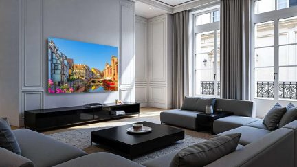 Salon contemporain lumineux, vue de face, avec canapé d’angle gris en tissu, table basse carrée noire en bois laqué, long meuble TV noir brillant portant un téléviseur mural à écran plat affichant une scène colorée, murs gris à moulures et grandes fenêtres à battants encadrées de rideaux gris et protégées par des garde-corps en fer forgé