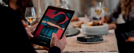 Tablet trzymany w dwóch rękach wyświetla kolorowy obraz, z nakrytym stołem i rozmytymi postaciami w tle.