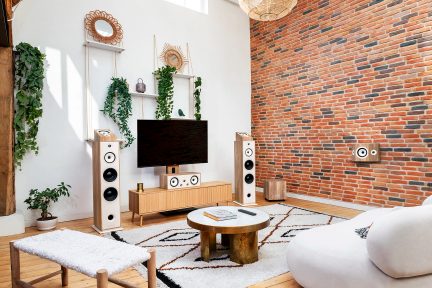 Un mur en briques, une télévision sur un meuble en bois, deux enceintes blanches, une table ronde, un tapis à motifs, des plantes suspendues
