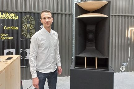 Vedere frontală a unei boxe acustice mari, dreptunghiulare, negre, din lemn, echipată cu două difuzoare concave bej suprapuse, un difuzor central circular și trei fante verticale, instalată într-un spațiu industrial lângă un bărbat îmbrăcat cu o cămașă deschisă la culoare