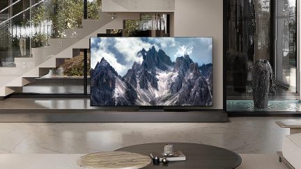 Téléviseur affichant une image de montagnes enneigées, posé sur un meuble dans une pièce avec de grandes baies vitrées