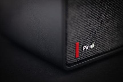 Vue en gros plan d’un haut-parleur rectangulaire noir, boîtier en plastique mat et façade en tissu maillé sombre, logo blanc « Pinell » rehaussé d’un l vert et d’une barre verticale rouge