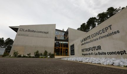 Clădire modernă din beton gri deschis, vedere frontală de jos în sus, fațade unghiulare decorate cu inscripții negre mari, intrare centrală vitrată cu montanți din lemn, rond de pietricele albe de-a lungul aleii asfaltate