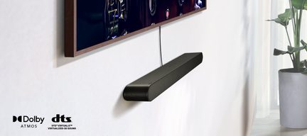 Rectangular black soundbar mounted on the wall beneath a wood-framed television, visible cable, potted plant on the right